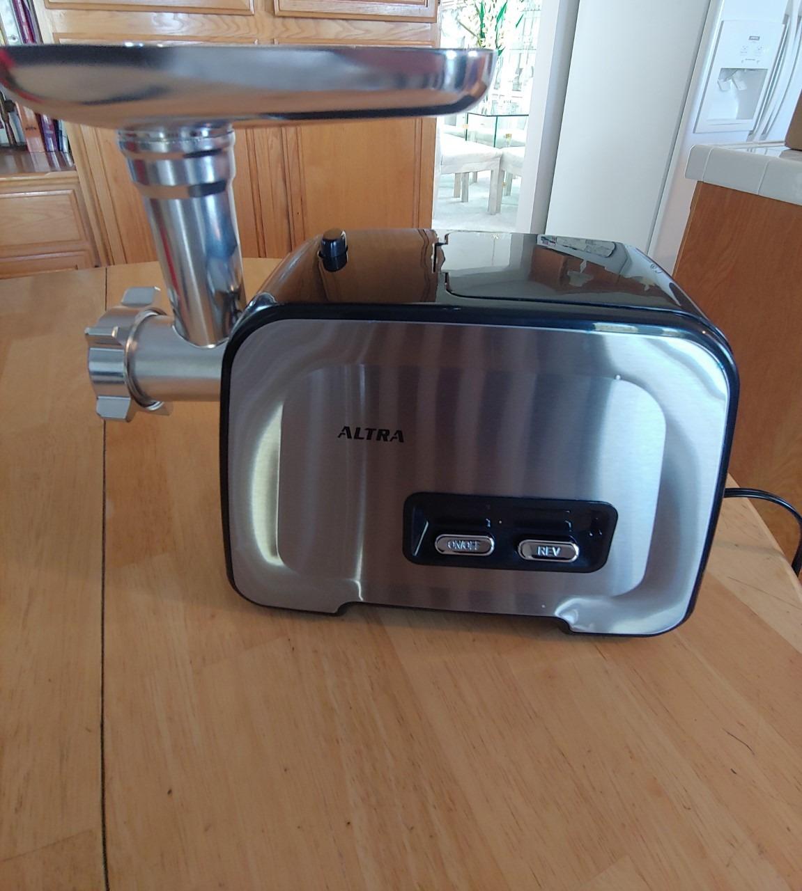Silver Altra meat grinder on a wooden kitchen table with the stainless tray fitted and the ON and REV buttons on the front panel