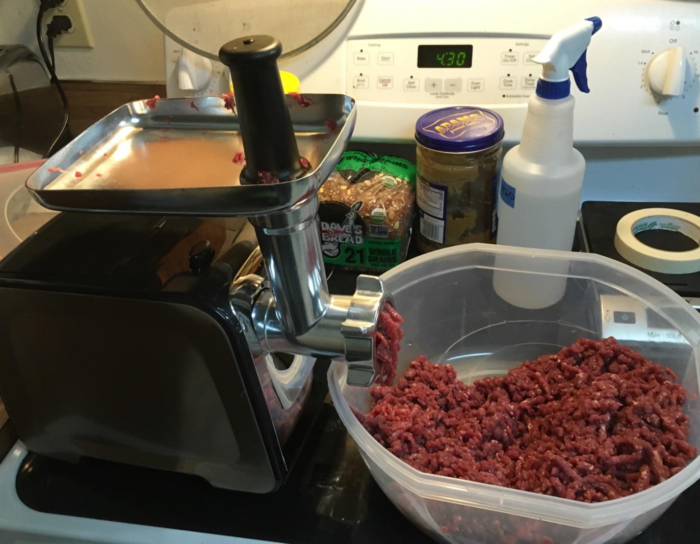 Altra meat grinder on a stovetop beside a large plastic tub filled with freshly ground red meat